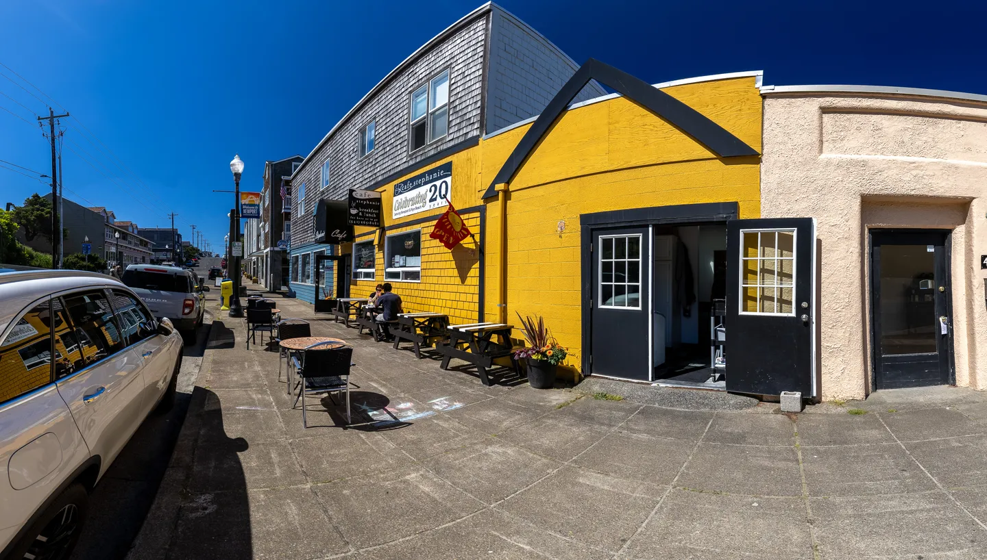 Street view showing a bright yellow storefront next to a cafe with outdoor seating along a sidewalk.