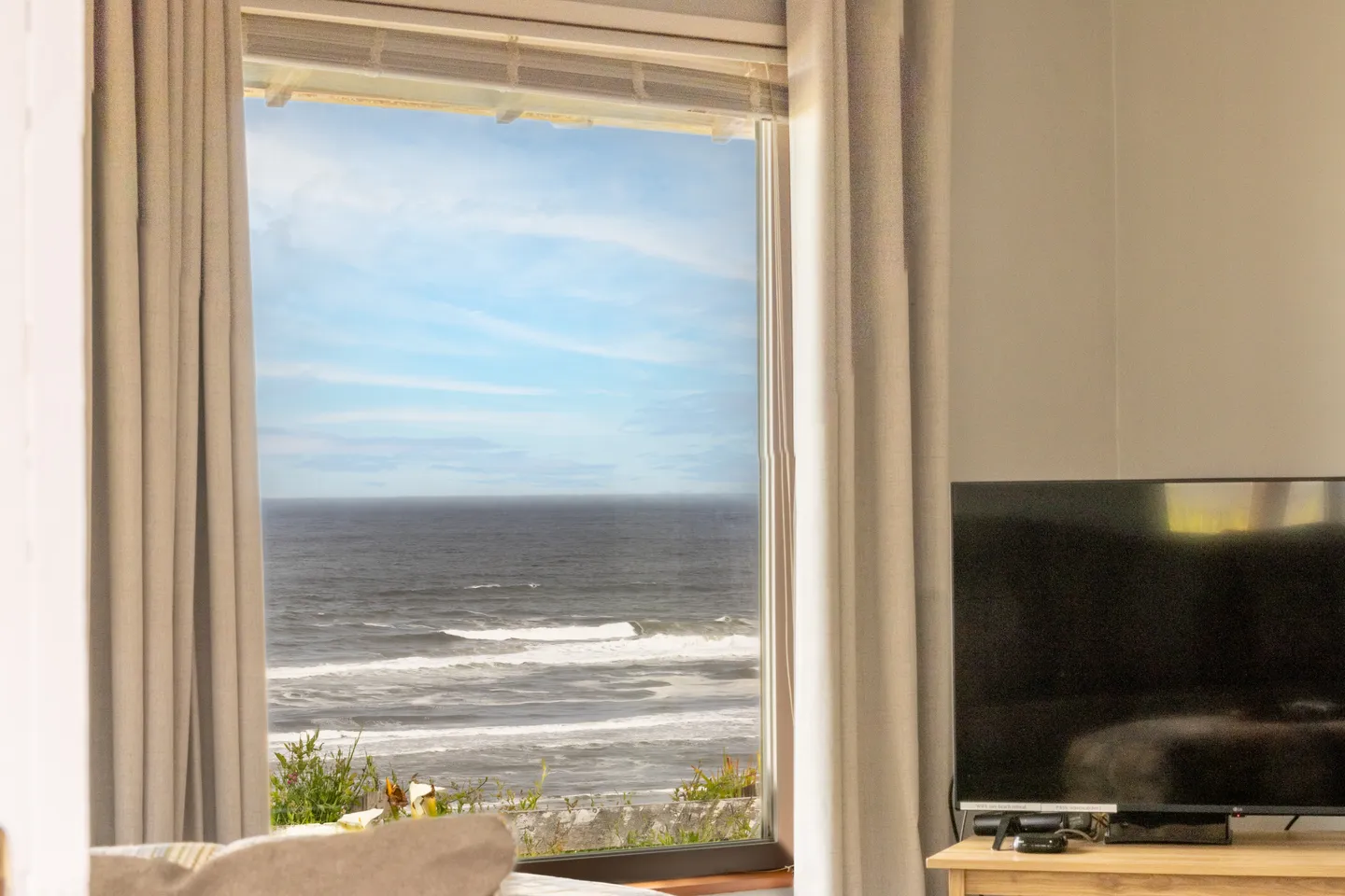 A large window looking out onto an expansive view of the ocean, flanked by curtains next to a television.