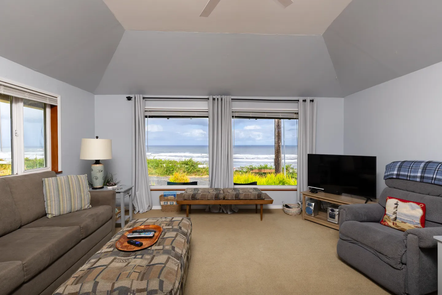 Living room with carpeted floors, vaulted ceiling, couch, recliner, and large windows looking out towards the ocean.