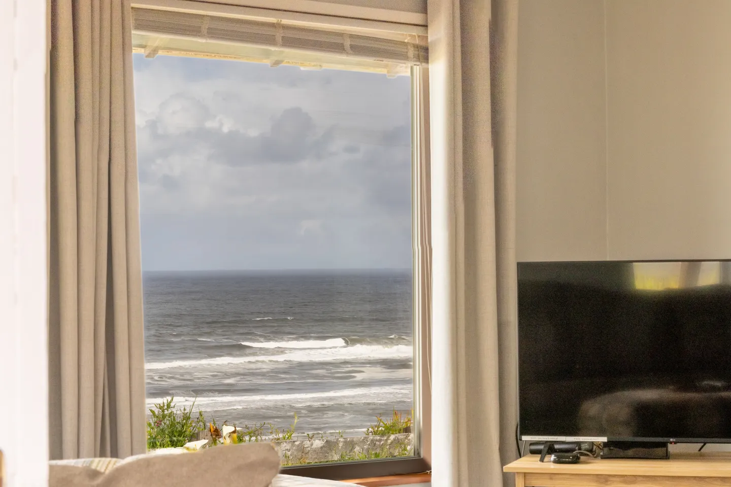 A large window framed by neutral curtains looking out at ocean waves under a cloudy sky.