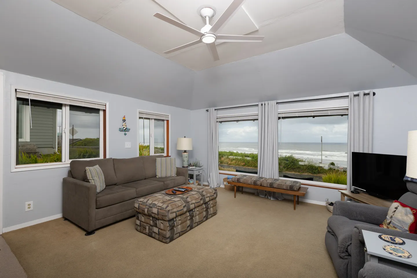 Living room with carpet, a brown sofa, patterned ottoman, armchair, and windows overlooking the ocean.