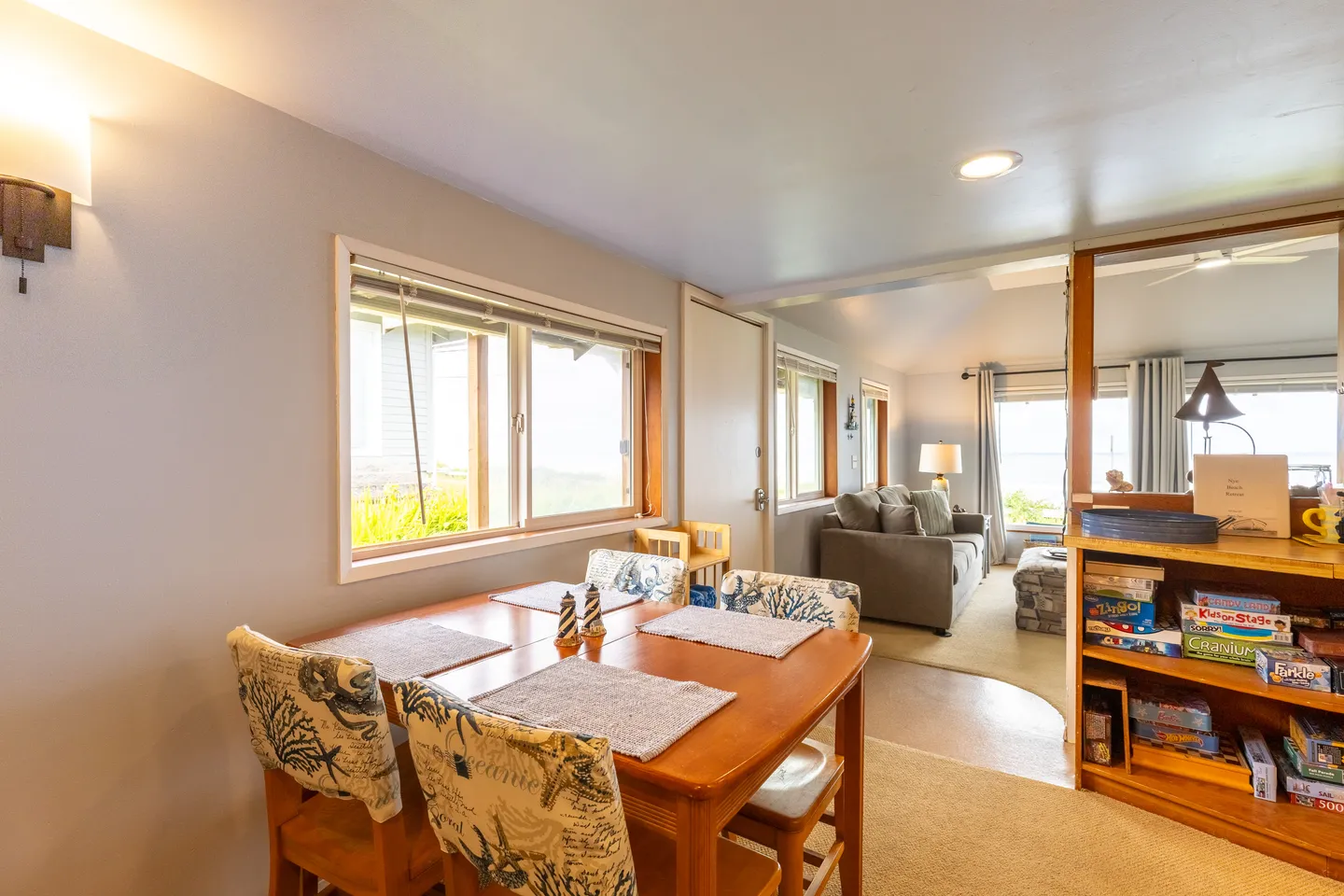 Dining area with a wooden table and patterned chairs, looking through to a living room with a sofa and windows.