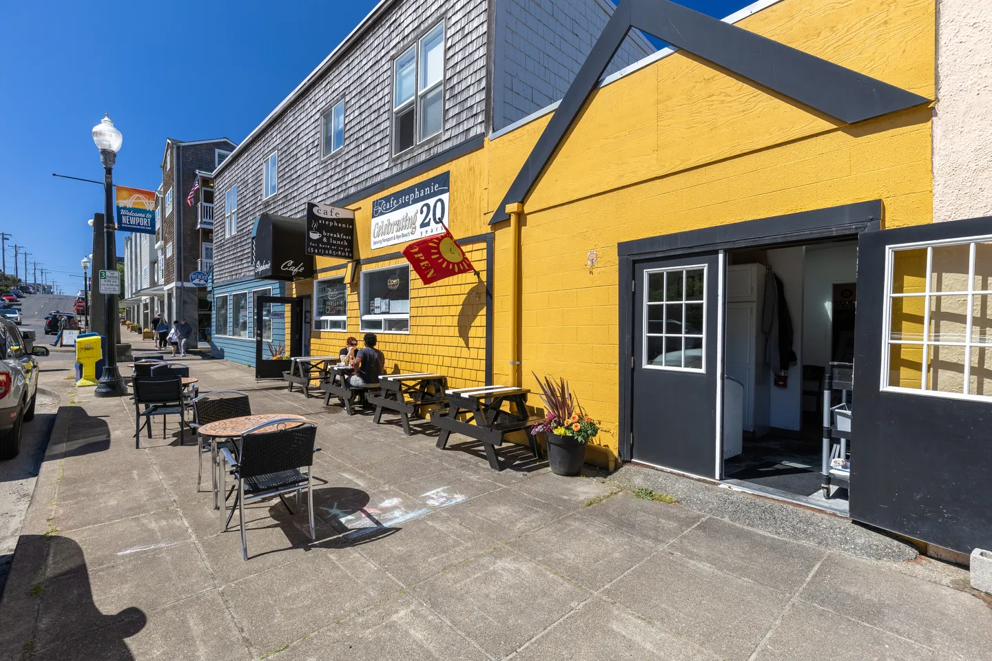 Street view of a yellow and wood-shingled building with outdoor cafe tables along the sidewalk on a sunny day.