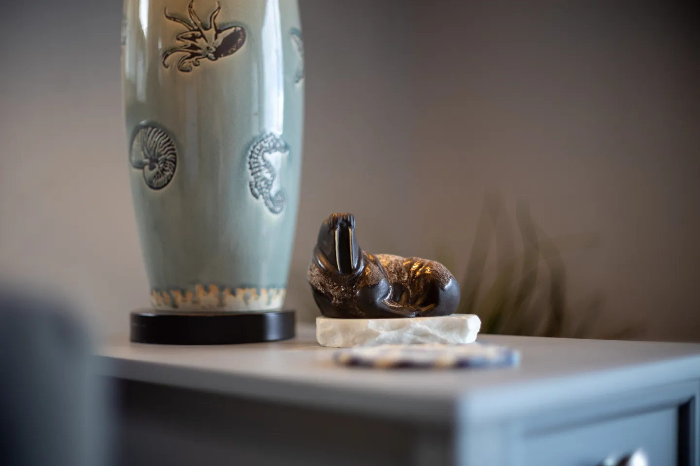 A close-up shot of a small walrus figurine resting on a stone base on a side table next to a ceramic lamp.