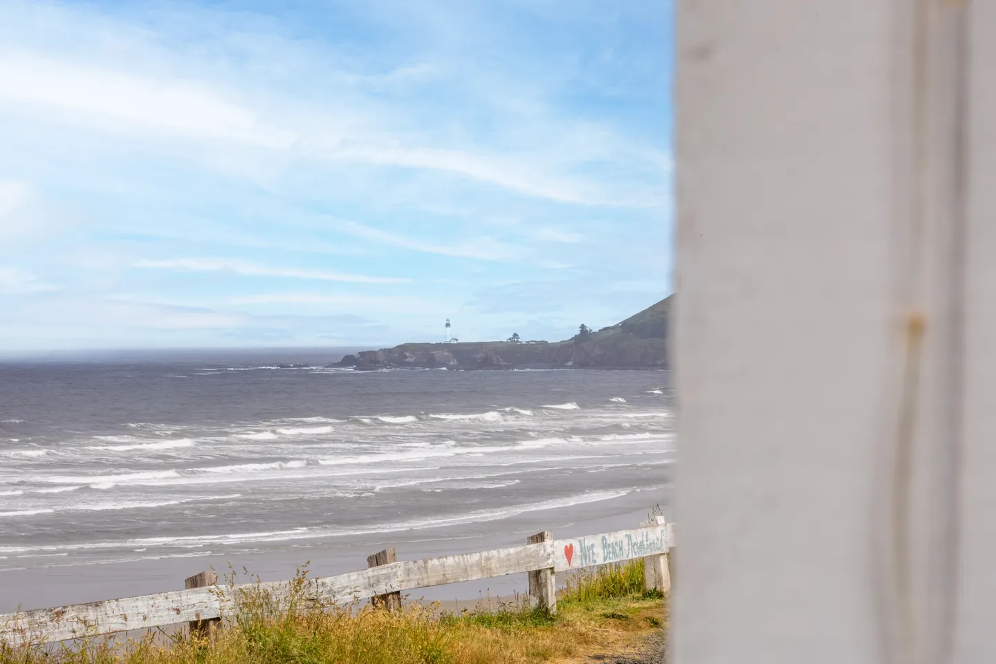 A coastal view of the ocean, sandy beach, and a distant headland framed by a white exterior wall.