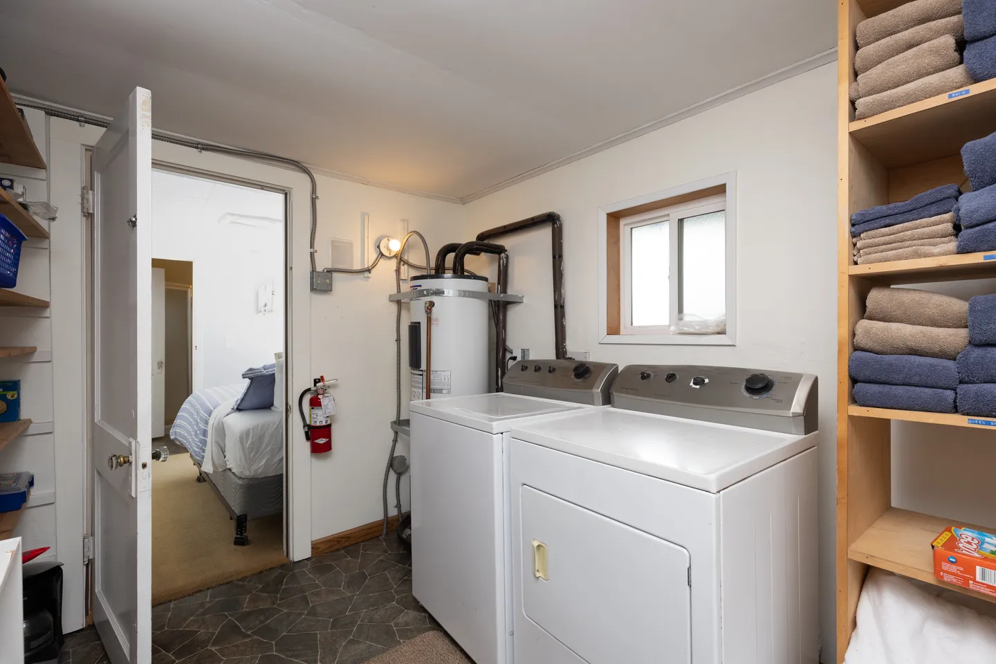 A utility and laundry room featuring a white washing machine and dryer, a water heater, shelves with folded towels, and an open doorway leading to a bedroom.