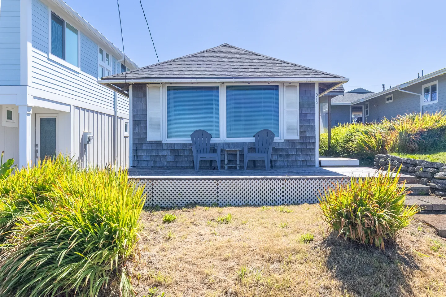 Exterior of a single-story wood shingle cottage featuring a front deck with two outdoor armchairs under a sunny blue sky.