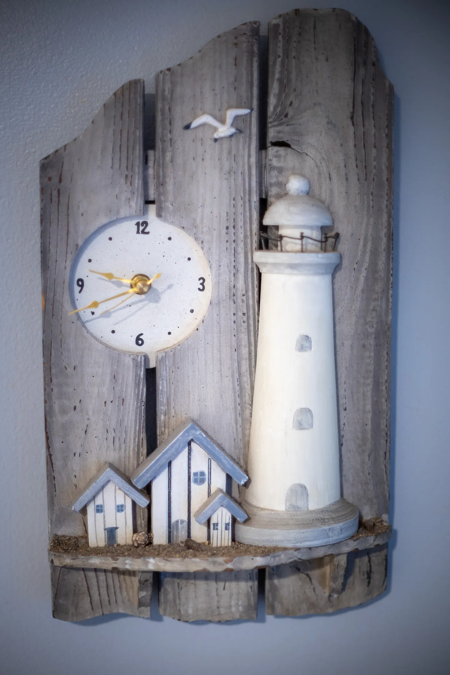 Nautical-themed wooden wall clock shaped like a lighthouse and coastal cottages mounted on a weathered wood plank base.