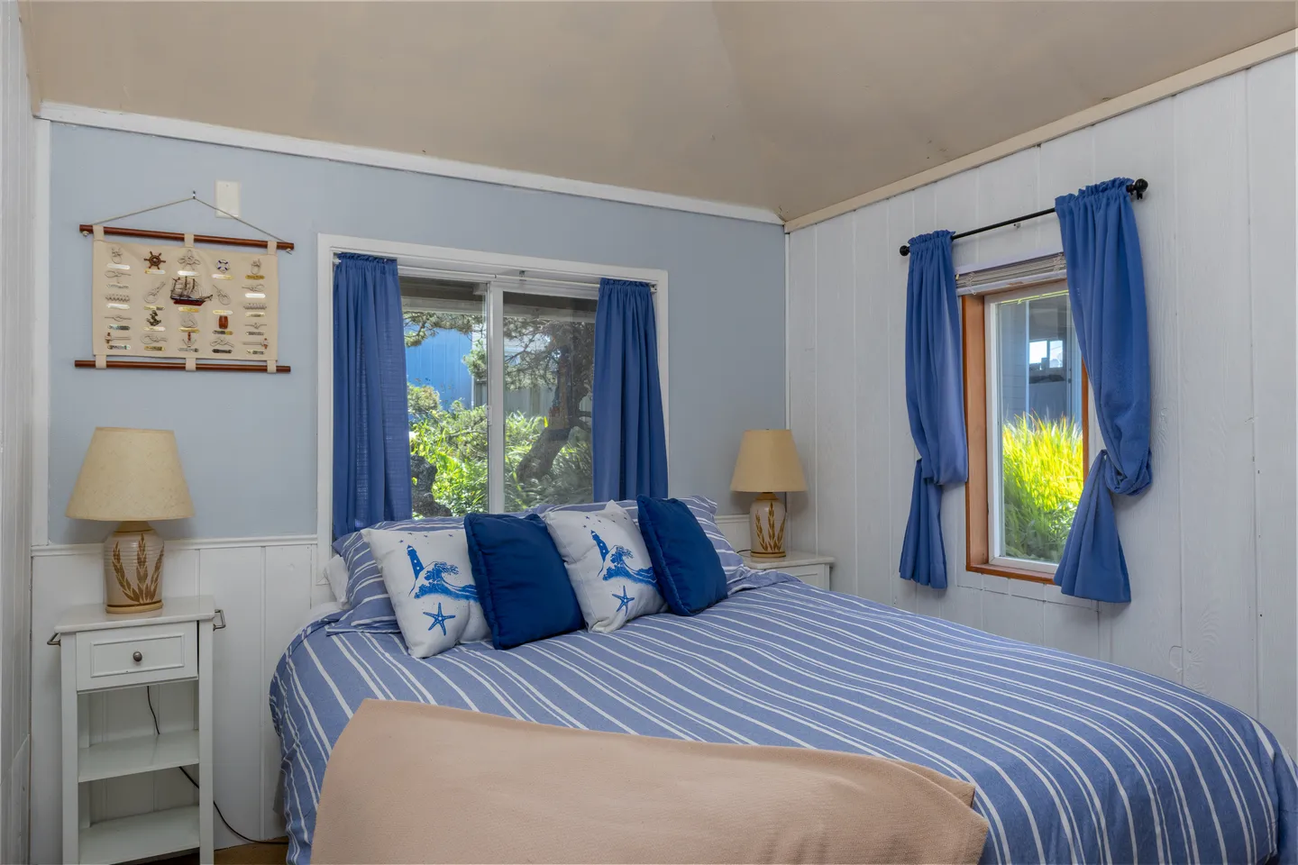 Bedroom with blue striped bedding, accent pillows, windows with blue curtains, and white paneled walls.