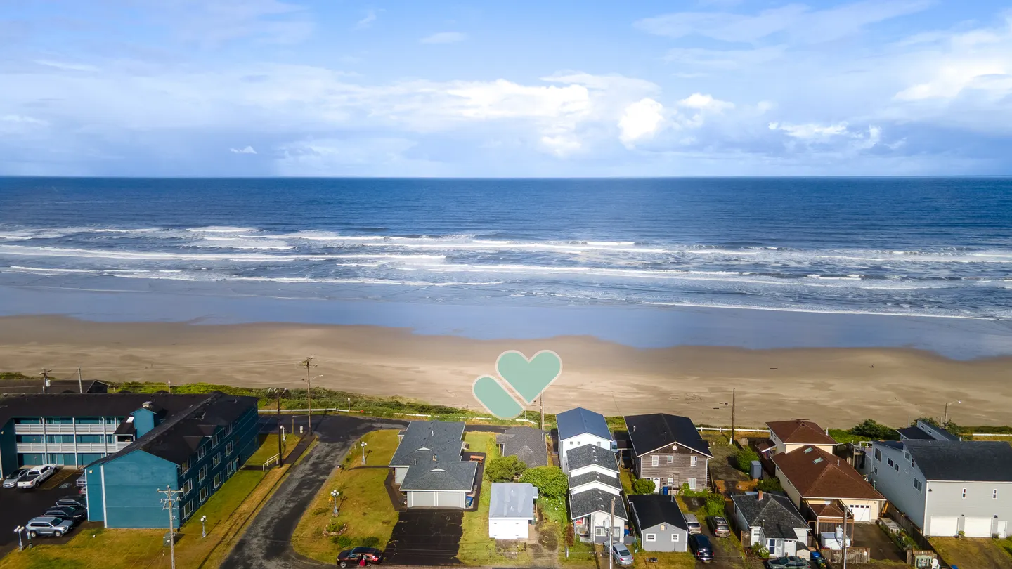 Aerial view of a coastal neighborhood with houses facing a wide sandy beach and ocean surf under a blue sky.