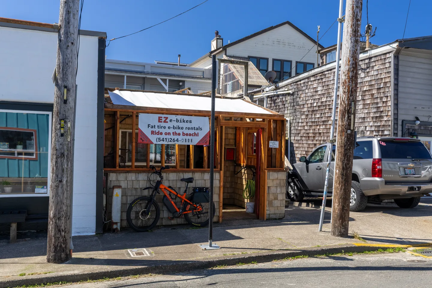 Exterior view of an electric bicycle rental shop along a street with parked vehicles and neighboring buildings under a clear blue sky.