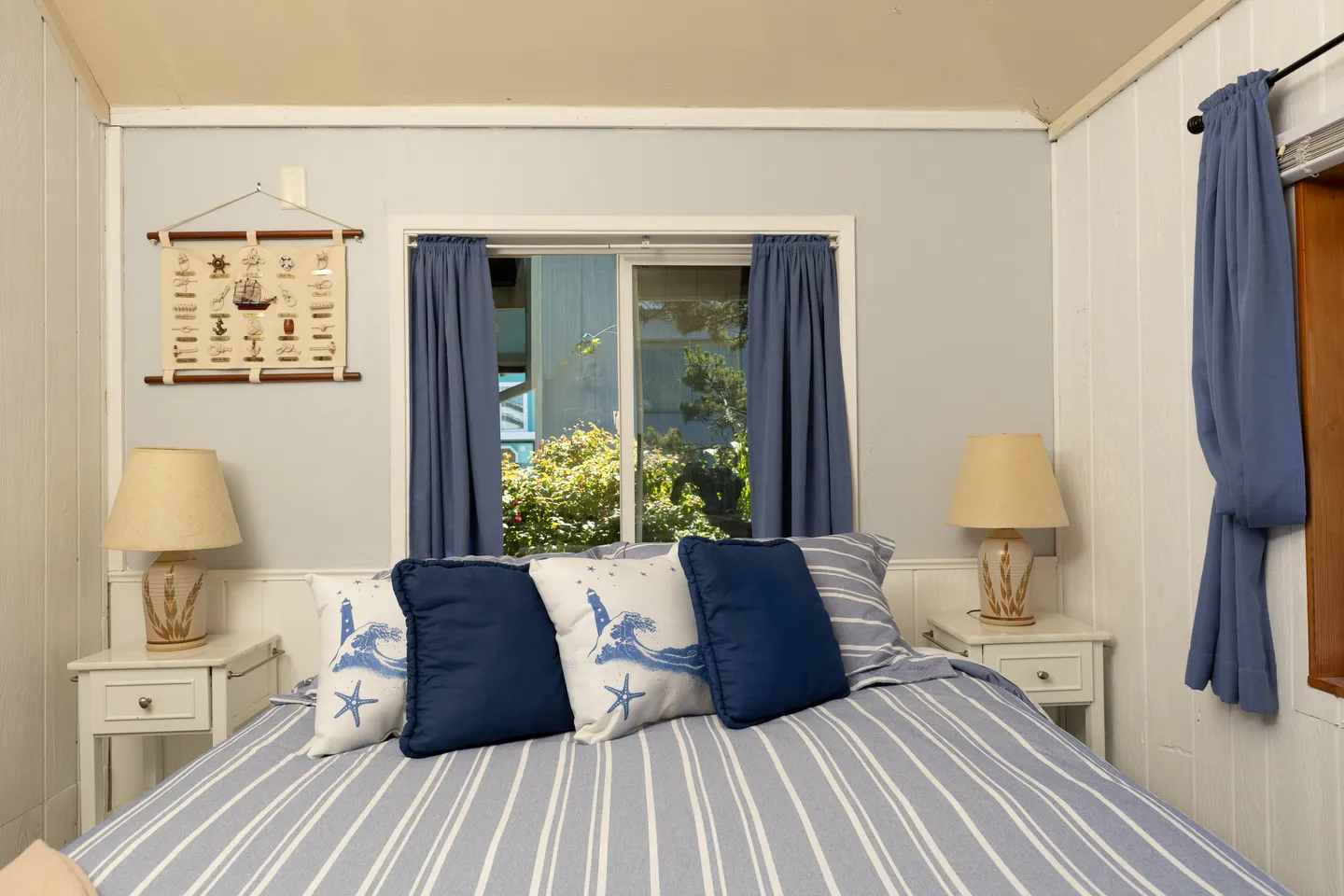 Bedroom with blue striped bedding, coastal accent pillows, a sliding window with blue curtains, and two nightstands with lamps.