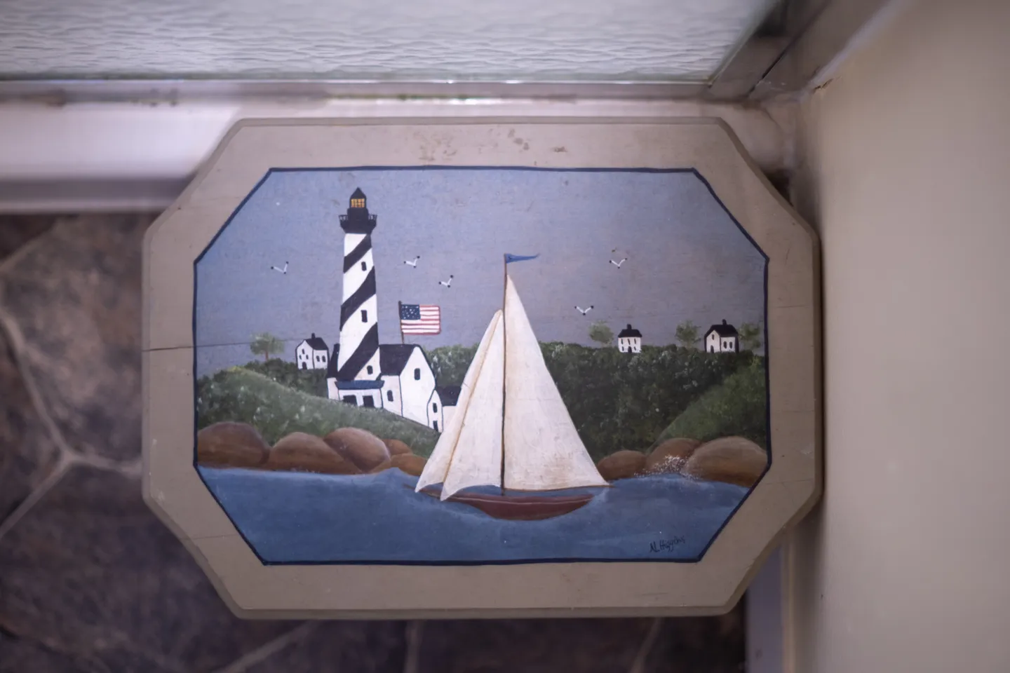 Decorative painted plaque depicting a sailboat, lighthouse, and coastal scene set on a textured surface.