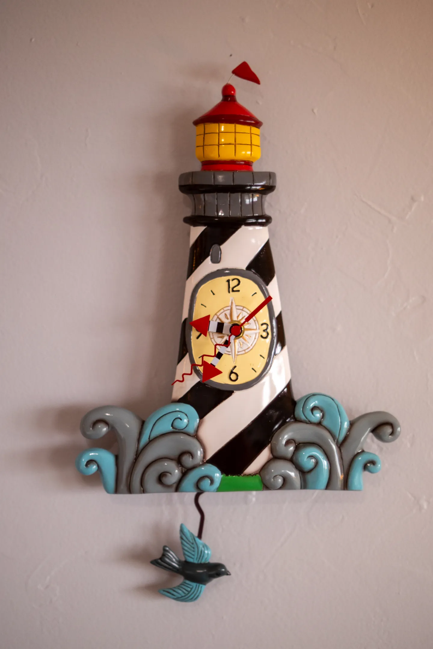 A decorative lighthouse-shaped wall clock with a bird pendulum hanging on a light-colored wall.