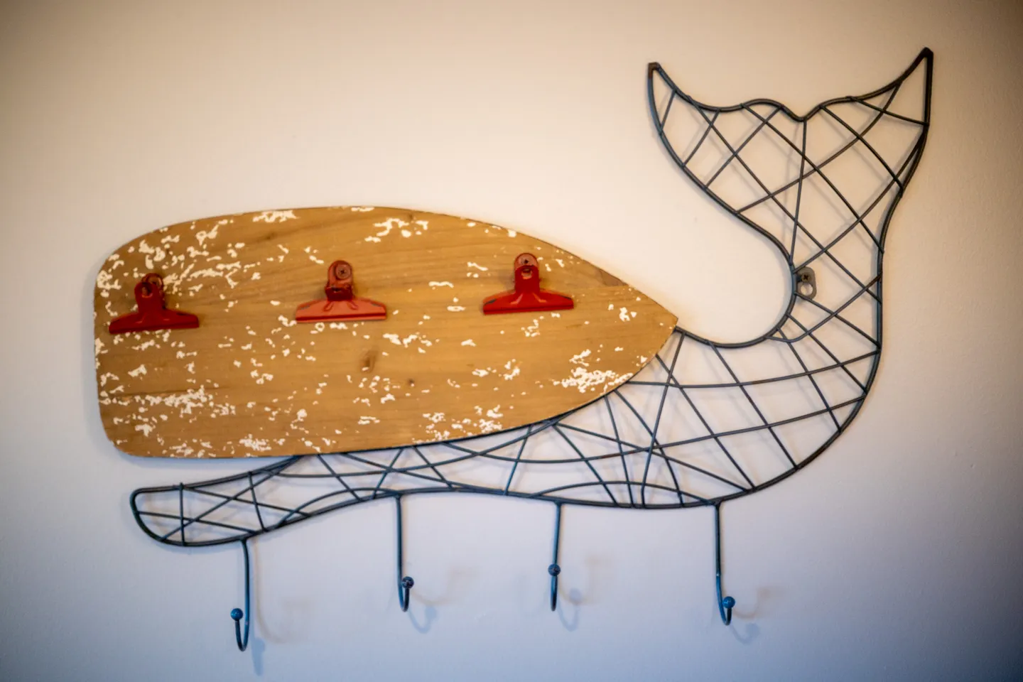 A whale-shaped wall hanger made of wire and distressed wood with three red clips and four hooks mounted on a plain wall.