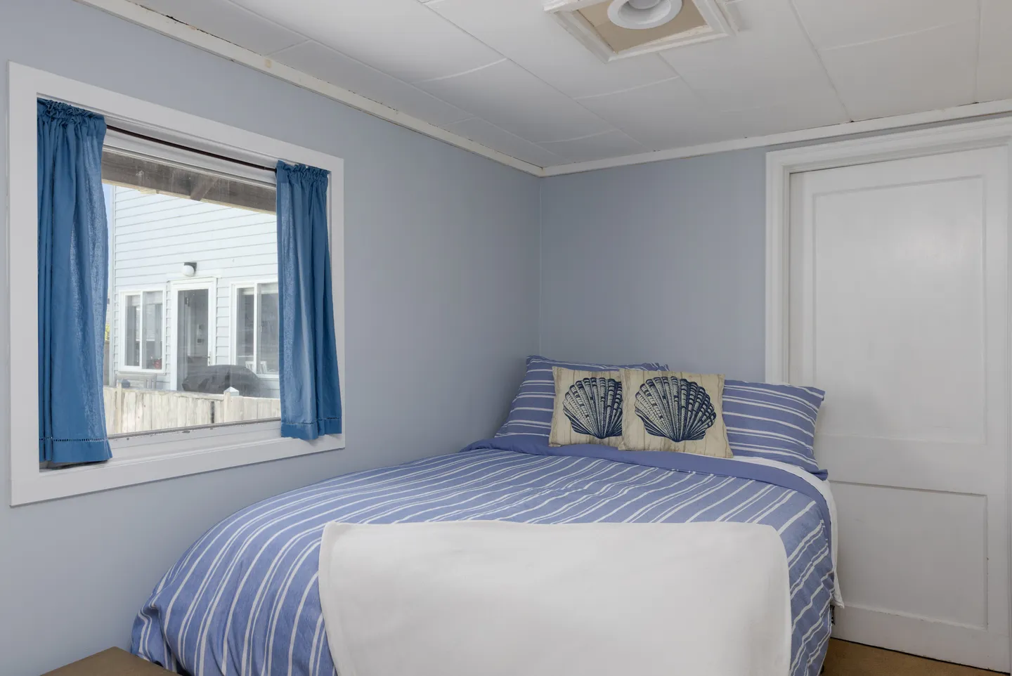 Bedroom with light blue walls, a bed dressed in blue and white striped bedding, and a window with blue curtains.