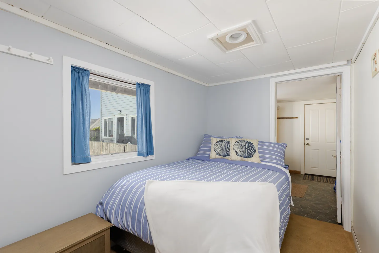 Bedroom featuring a made bed with blue and white striped bedding, seashell accent pillows, a window with blue curtains, and a doorway leading to an adjoining room.