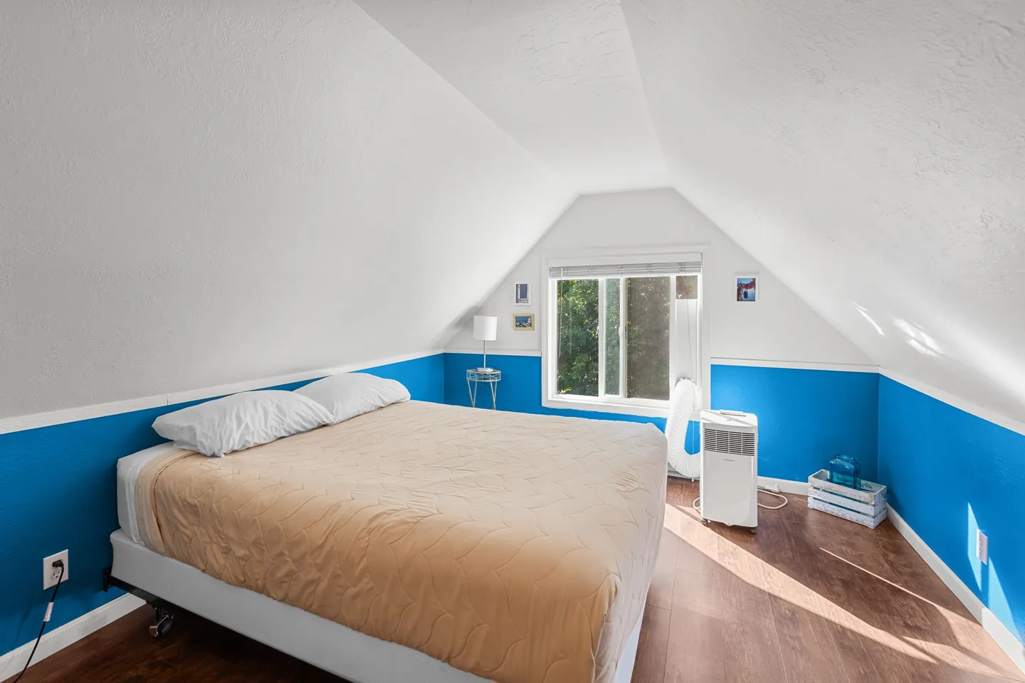 Attic bedroom with sloped ceilings, vibrant blue lower walls, a bed made with a tan coverlet, and wood-look flooring.