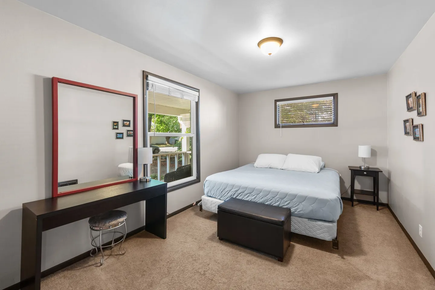 Bedroom with carpet flooring, a bed, a vanity table with a red-framed mirror, and two windows.