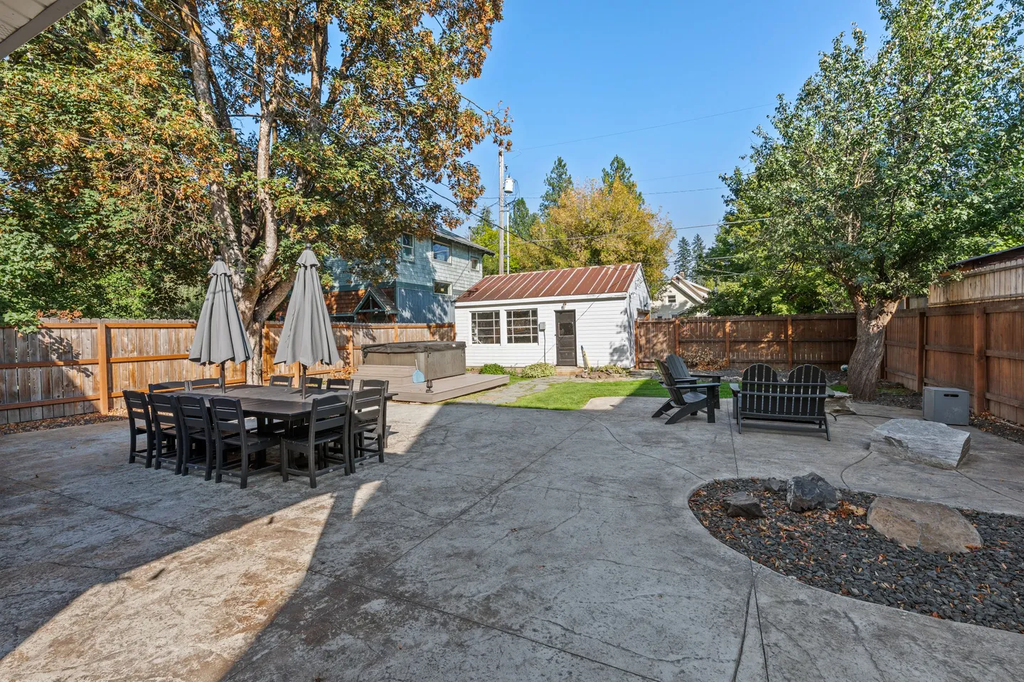 Fenced backyard patio featuring a large outdoor dining table, a hot tub, a fire pit area, and a detached outbuilding.