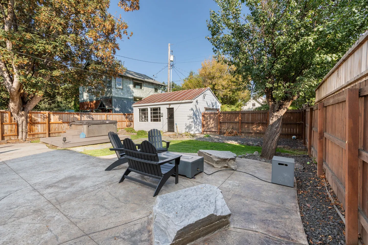 Fenced backyard patio with outdoor seating, a fire pit, large boulders, a hot tub on a deck platform, and a detached outbuilding under mature trees.