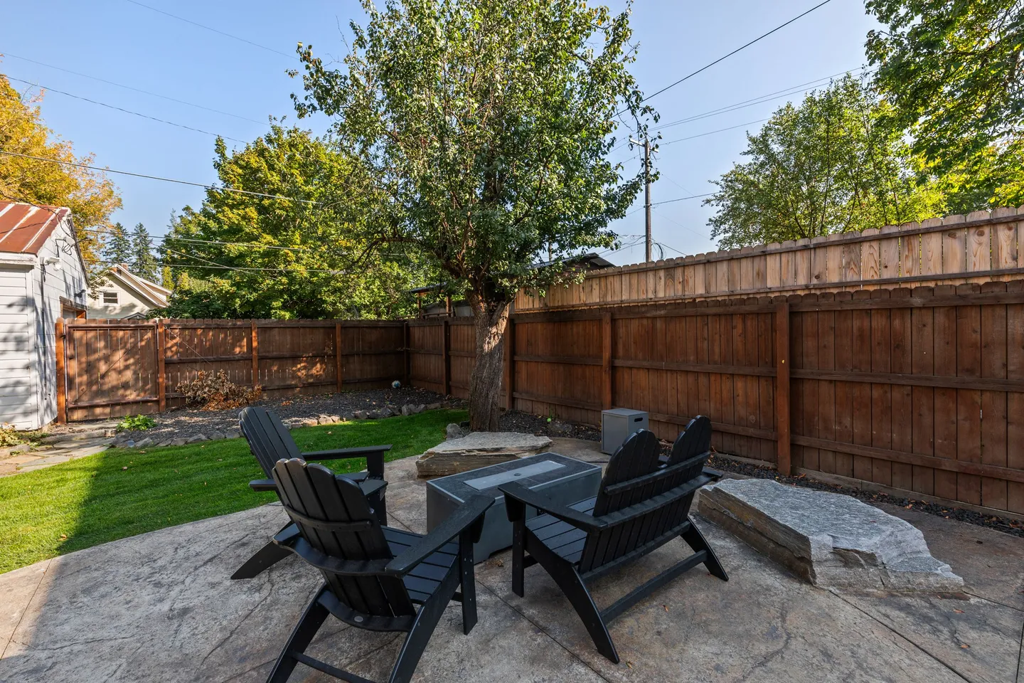 Fenced backyard patio featuring dark Adirondack chairs around a central fire table with grass and a tree in the background.