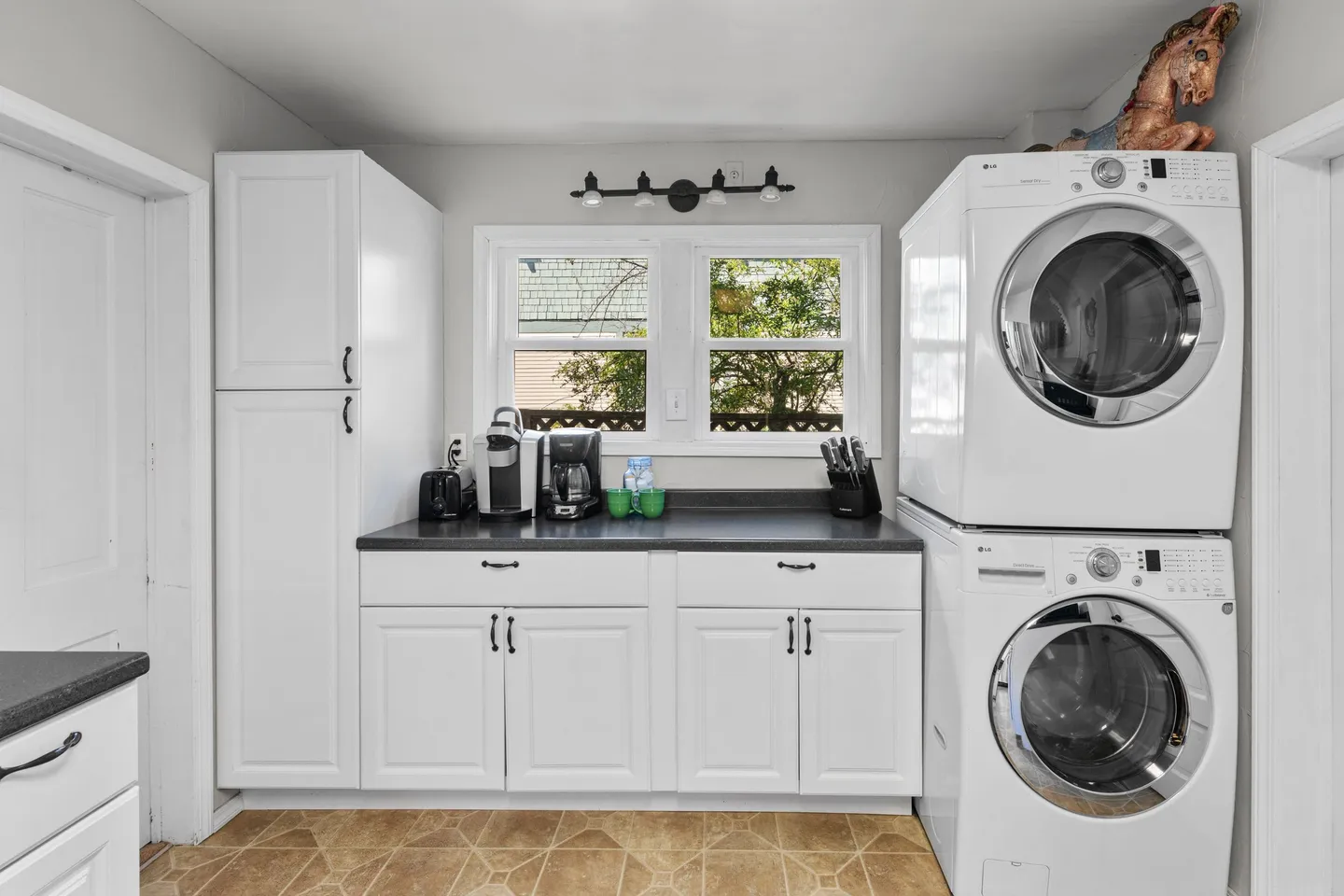 Laundry and coffee nook with white stacked washer and dryer, white cabinets with dark countertops, coffee makers, and a window.