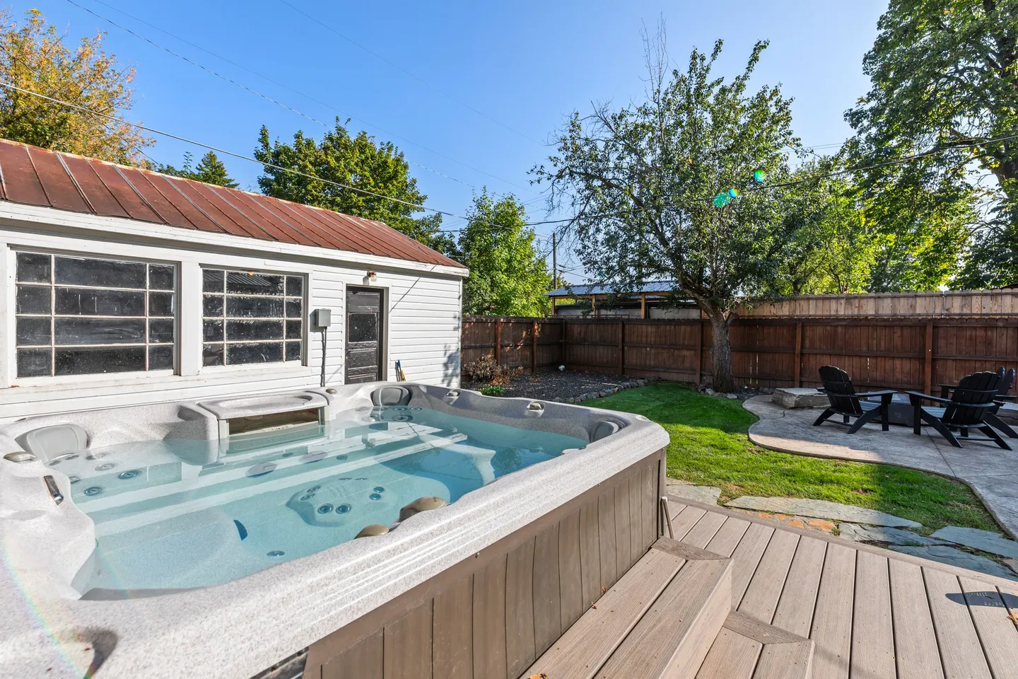 Outdoor hot tub on a wooden deck in a fenced backyard next to a white outbuilding and fire pit area.