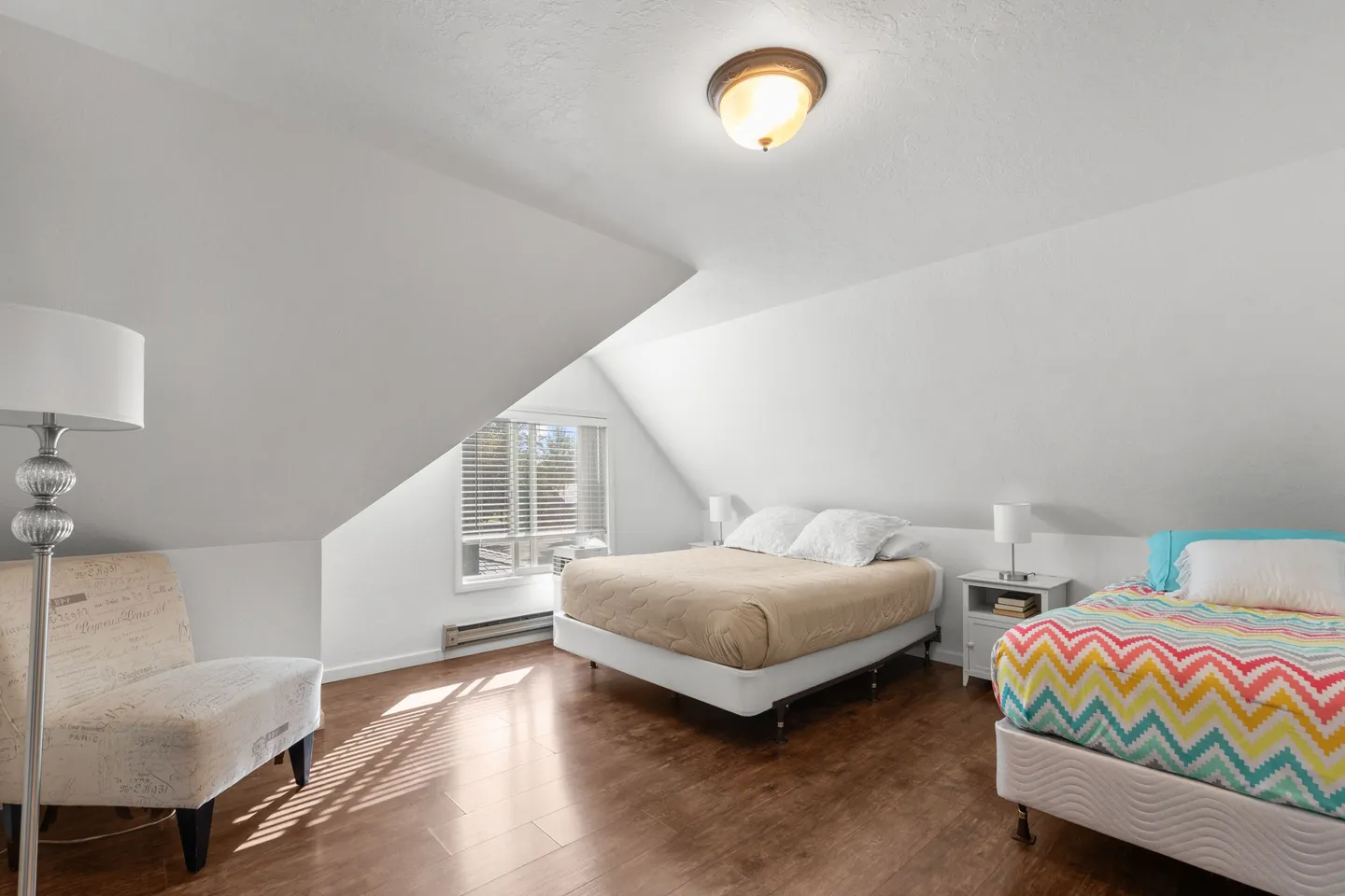 Bedroom with sloped ceiling, wood-look flooring, two beds, an armchair, and a window with blinds.