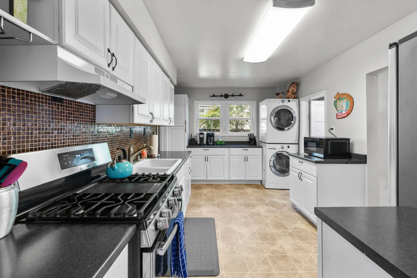 Kitchen with white cabinetry, black countertops, gas range, mosaic tile backsplash, and stacked washer-dryer unit.