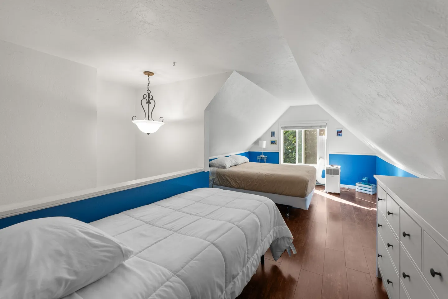 Upstairs loft bedroom with slanted ceilings, two beds, white and blue walls, and a window at the end.