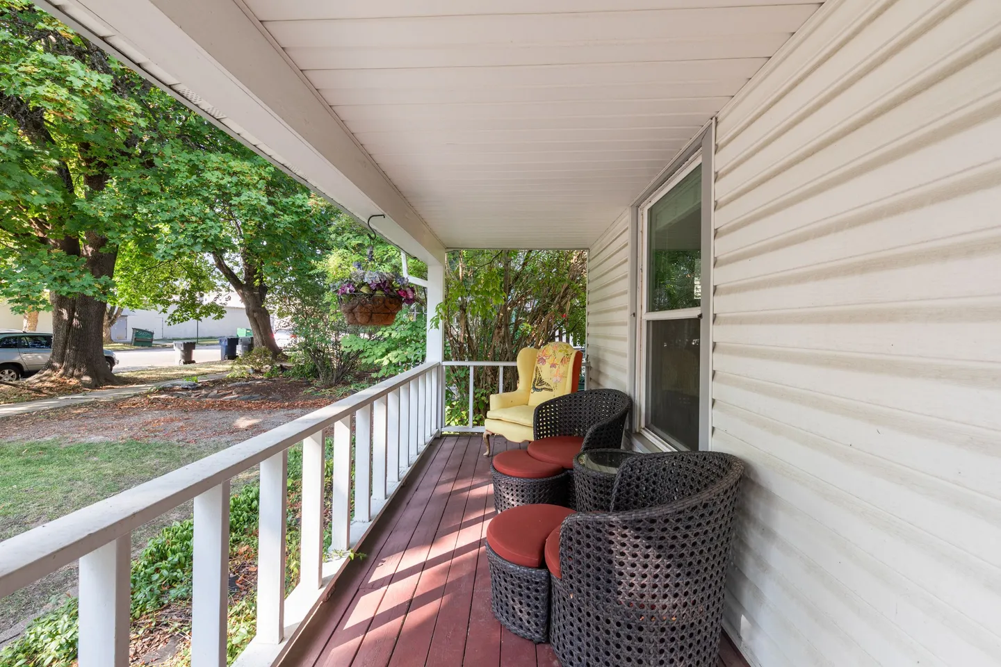 Covered front porch with white railing, woven outdoor armchairs with red footstools, a yellow accent chair, and a view of the street.