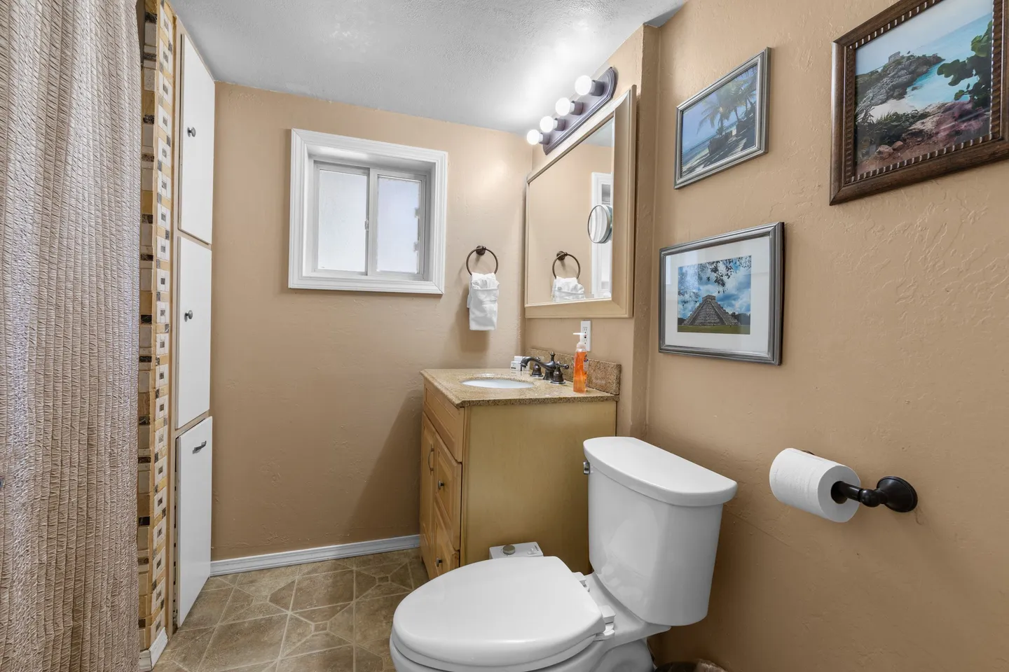 Bathroom with a toilet, vanity sink with mirror and vanity lights, a small frosted window, and framed art on warm tan walls.