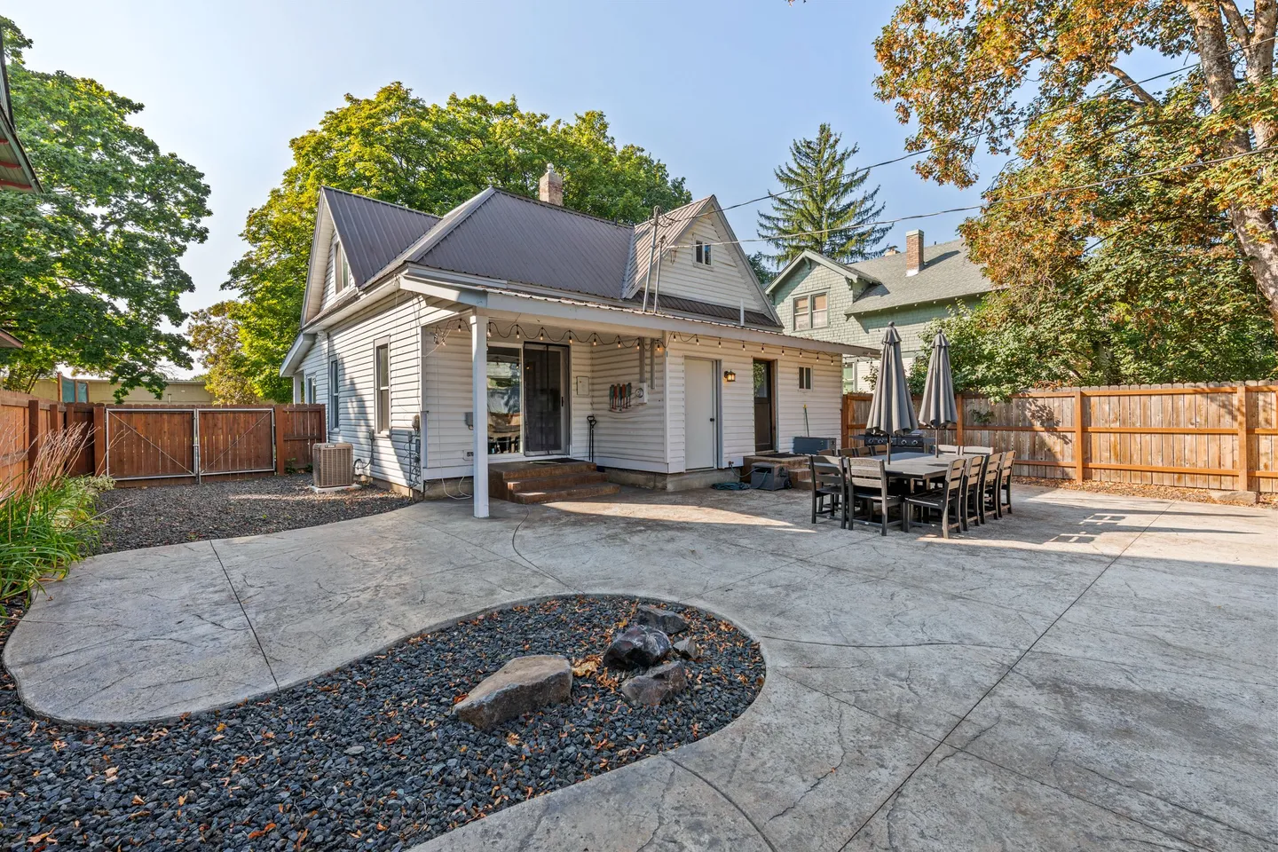 Fenced backyard patio behind a house, featuring an outdoor dining table, string lights, and a small rock feature.