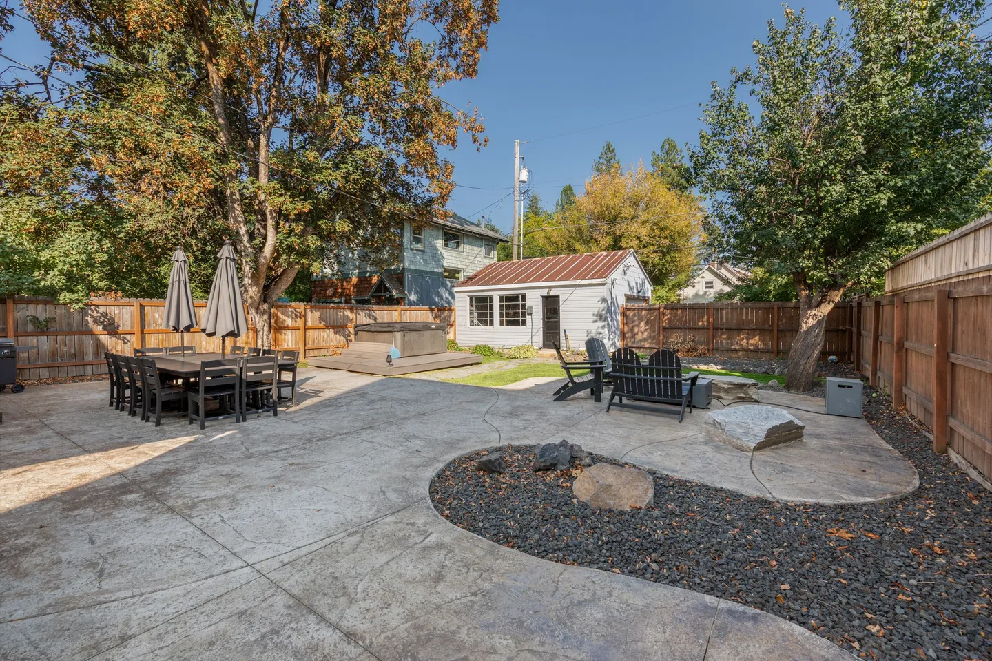Fenced backyard patio featuring a dining set with umbrellas, Adirondack chairs around a fire feature, and a hot tub on a platform.