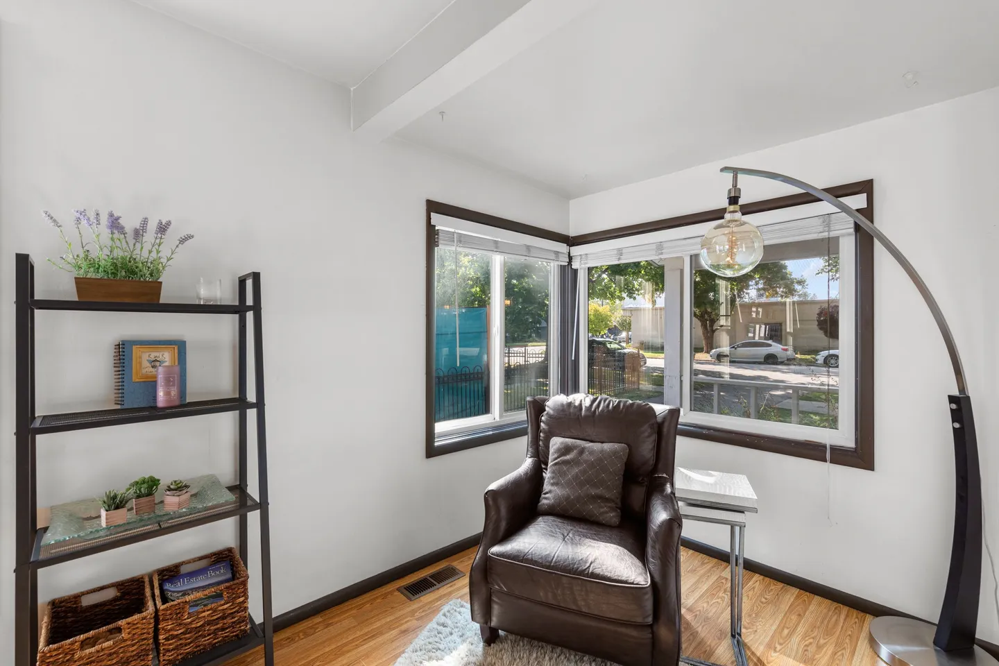 A reading corner with a brown leather armchair, arched floor lamp, open shelving unit, and corner windows looking outside.