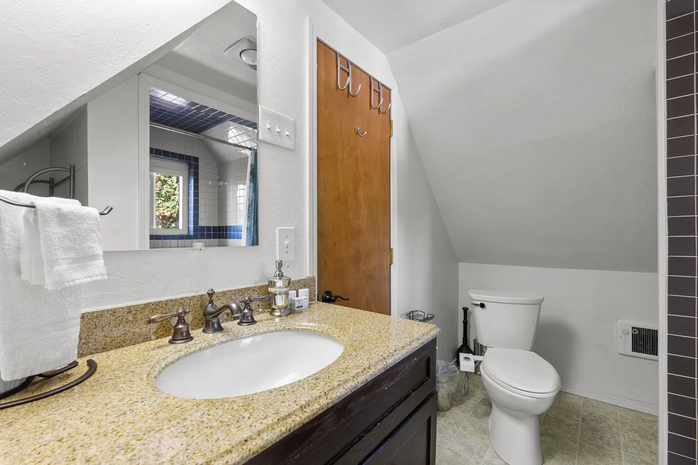 Bathroom with sloped ceiling, vanity with speckled countertop, mirror reflecting a tiled shower, and a white toilet.