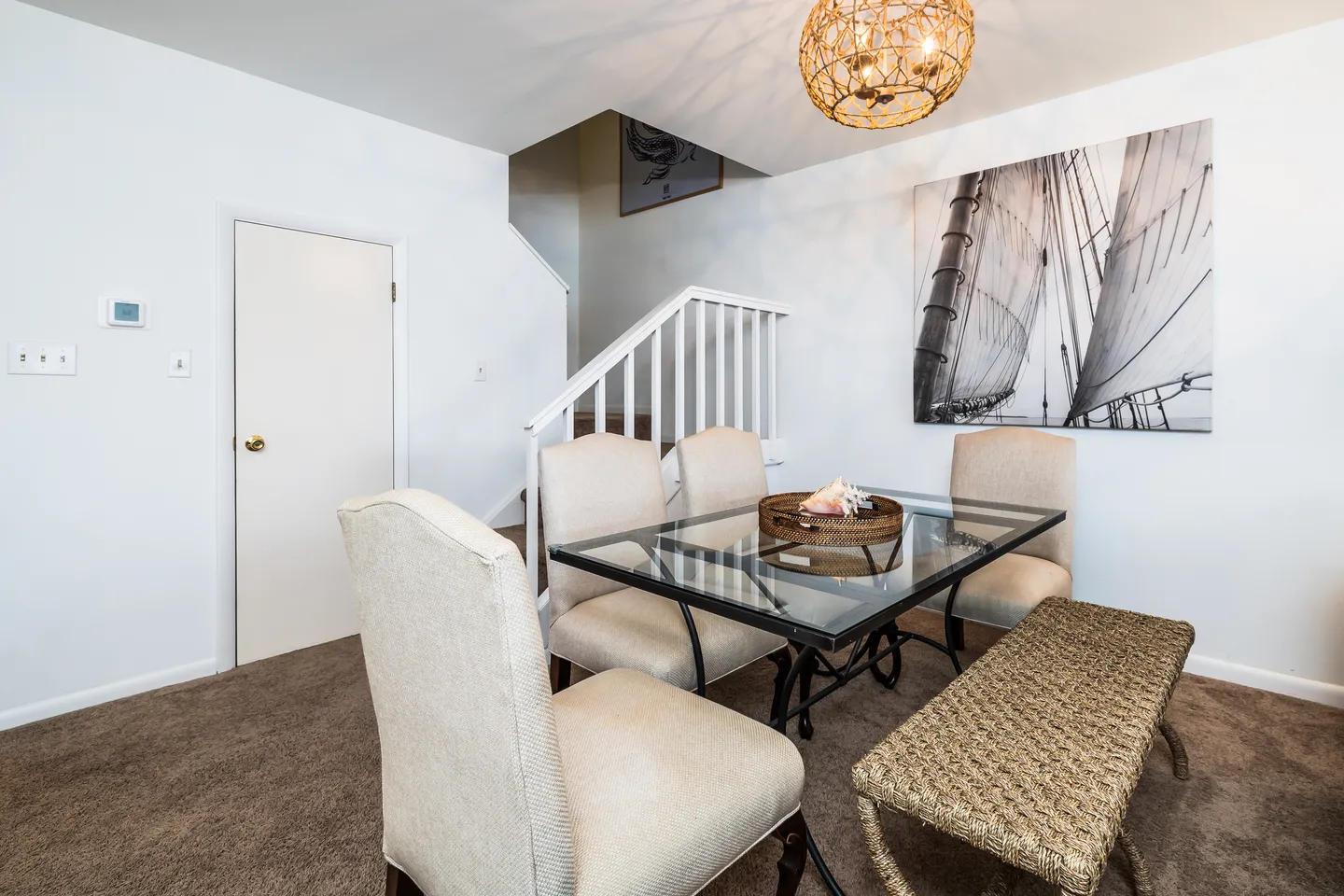 Dining area with a glass-top table, upholstered chairs, a woven bench, and a staircase in the background.