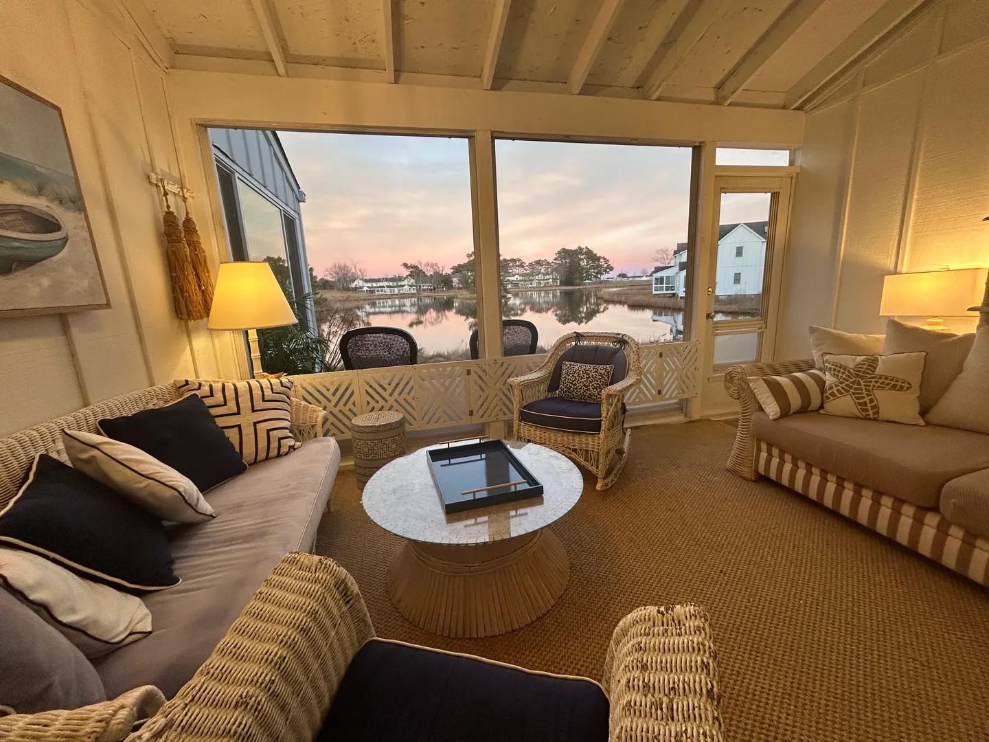 Screened porch furnished with wicker sofas, armchairs, and a coffee table, overlooking a calm body of water at dusk.