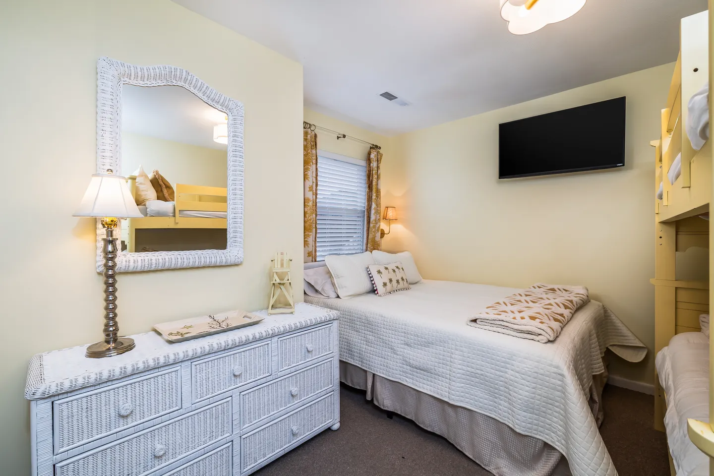 Bedroom with a white-dressed bed, bunk beds, a white wicker dresser with a mirror, and a wall-mounted TV.