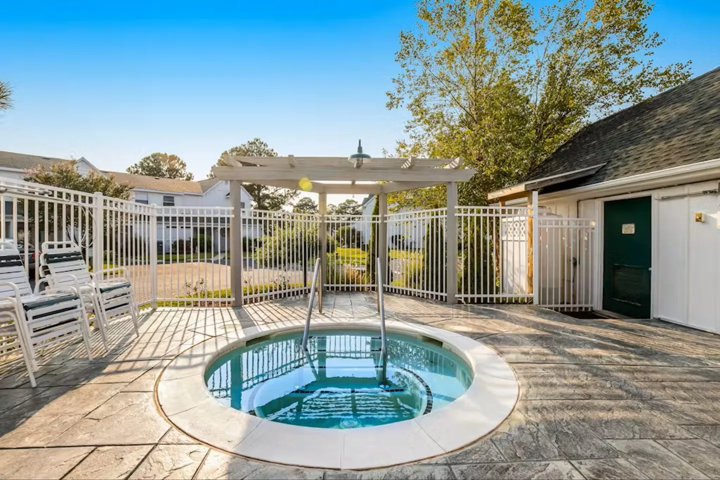 Circular outdoor hot tub set into a paved patio surrounded by a white metal fence with lounge chairs nearby.