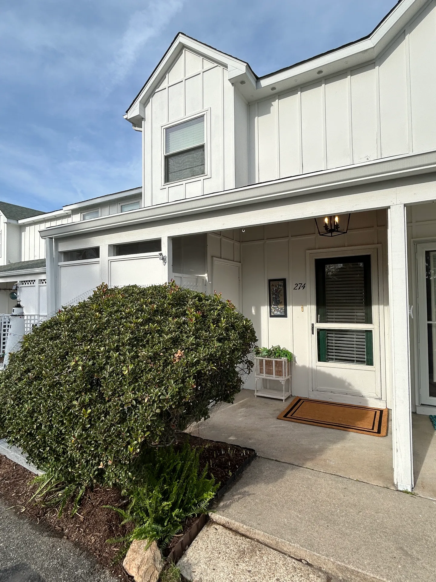 Front entrance of a white two-story townhome with a covered porch, green front door, and adjacent large bush.