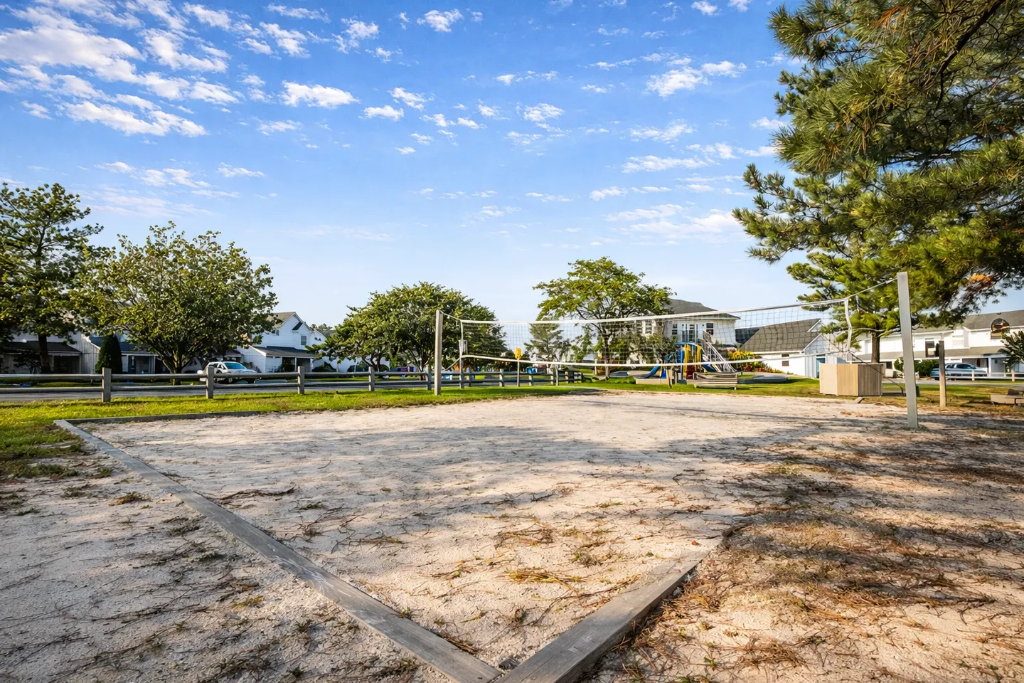 Outdoor sand volleyball court with net under a blue sky surrounded by trees and nearby houses.