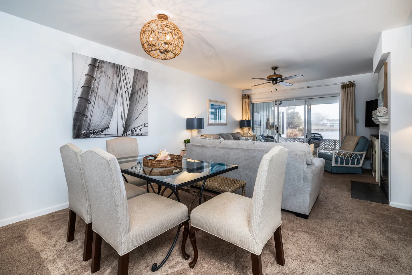 Dining area with a glass-topped table and upholstered chairs opening up to a living room with a sofa and sliding glass doors.