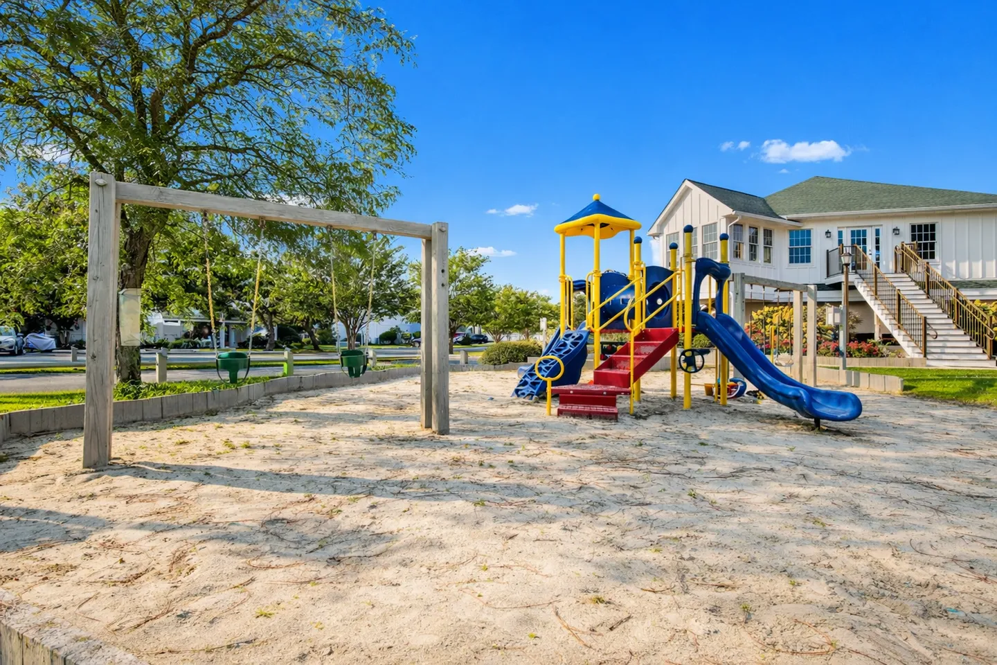 Outdoor playground area with a swing set and a blue and yellow play structure with slides set in a sand pit beneath a clear blue sky.