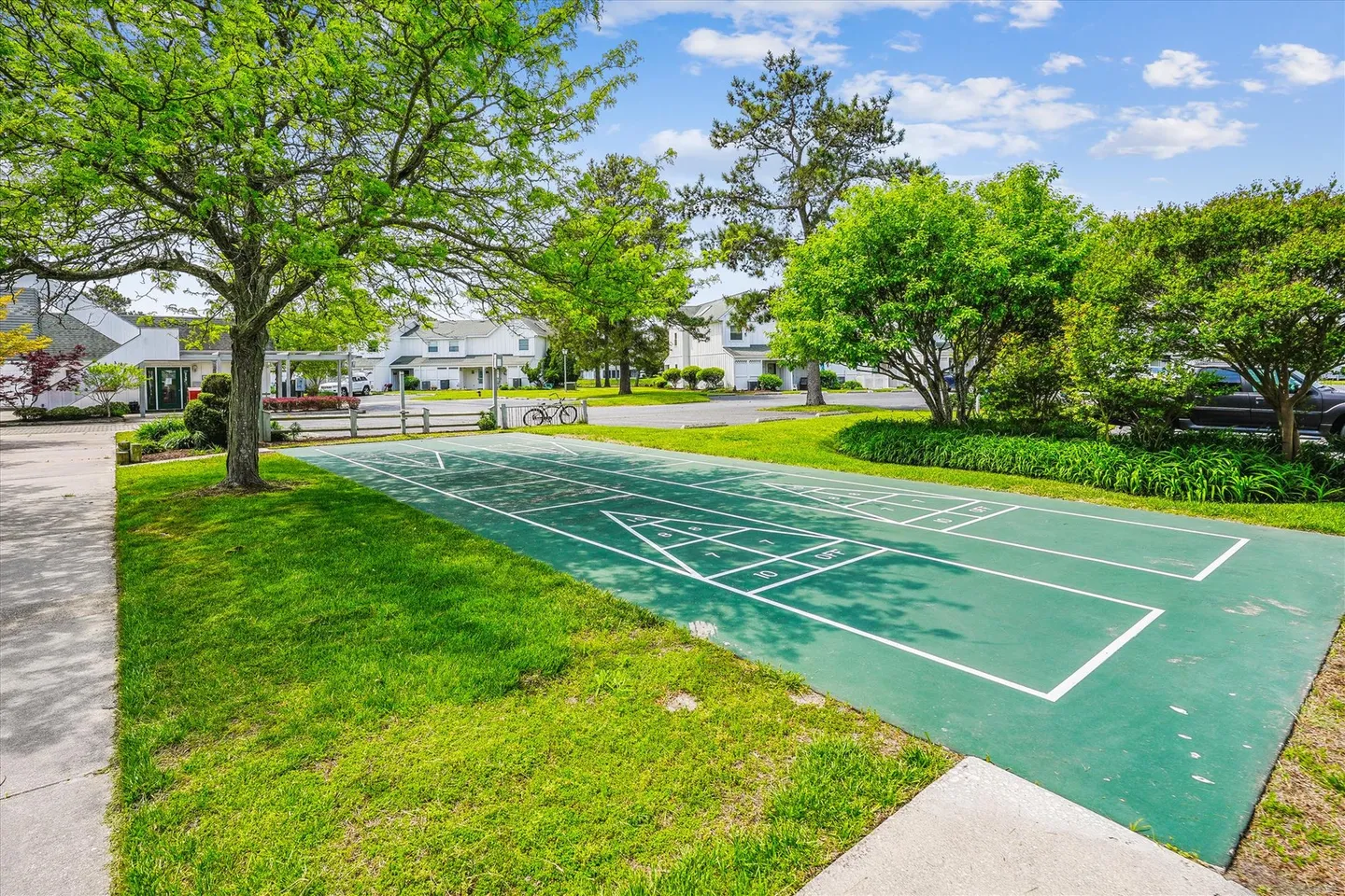 Outdoor green shuffleboard court bordered by grass, trees, and community buildings under a blue sky.