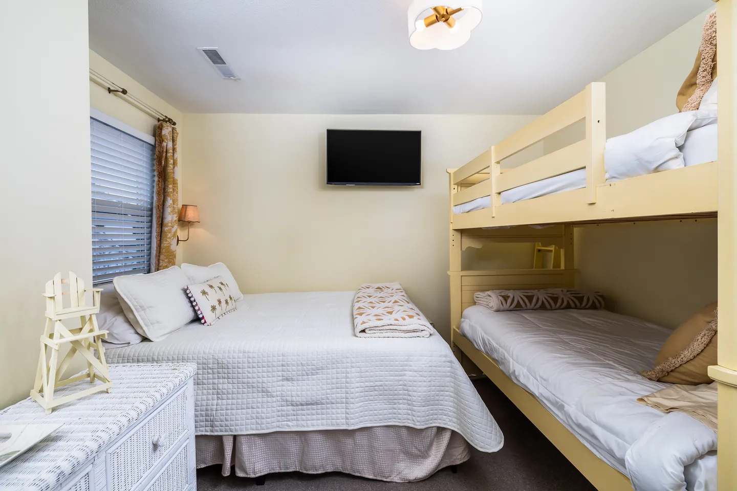 Bedroom featuring a double bed alongside a light-colored wooden bunk bed, with a wall-mounted TV and a white wicker dresser.