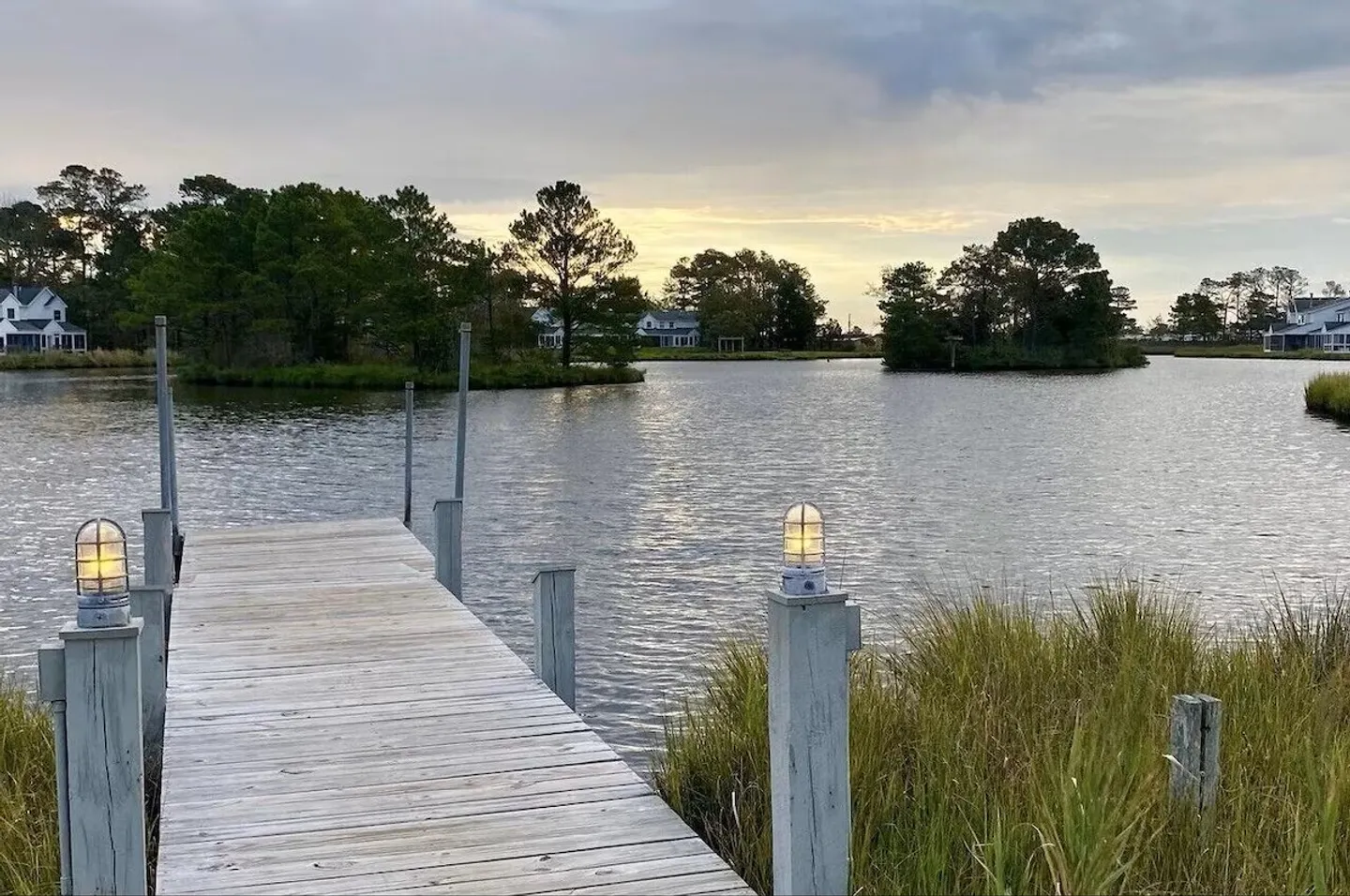 A wooden dock flanked by lit post lamps extends into a calm body of water with tree-lined shores in the background.