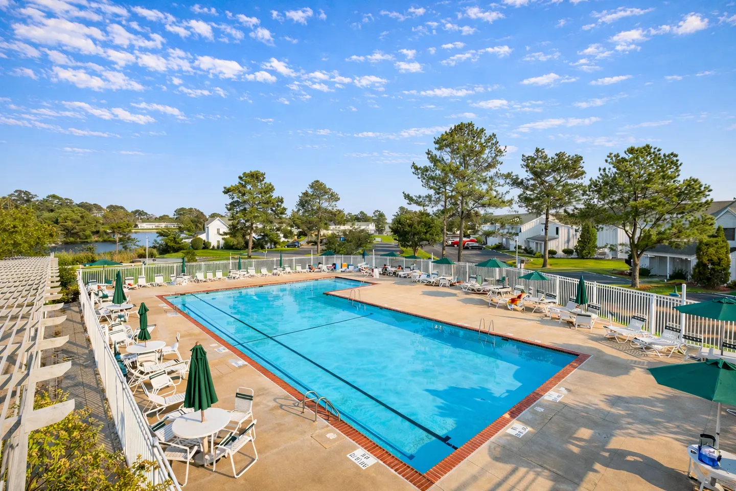 Elevated view of a large outdoor swimming pool surrounded by a concrete deck with lounge chairs, tables, and green umbrellas enclosed by a white fence.
