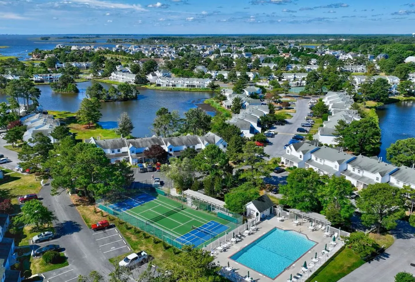 Aerial view of a coastal residential community featuring an outdoor swimming pool, tennis and pickleball courts, and a surrounding lake.